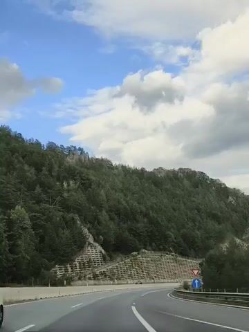 A white car drives on a highway. The car is on the left lane. The highway curves to the right. There is a mountain on the left side of the highway. The mountain is covered with trees. There are some concrete structures on the mountain. The sky is blue with white clouds.