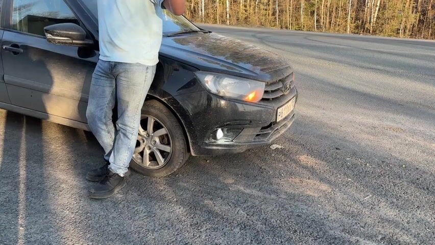 A man leans against the driver's side of a dark-colored car parked on the shoulder of a road. He wears light blue jeans, a light blue T-shirt, and dark shoes. The car's right turn signal is blinking. The road is paved and shows signs of wear. Trees line the road in the background.