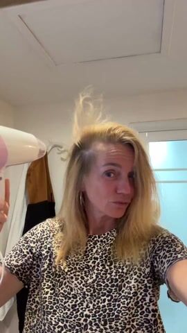 A woman dries her hair with a hairdryer in a brightly lit bathroom. She is wearing a leopard print top and has blonde hair, which is partially up in a messy style. In her left hand, she holds a small, light-colored brush as she directs the hairdryer, which is pink and white, towards her hair. The background shows a white door with a blue frame, a white towel hanging on a rack, and a beige jacket. There is also a white ceiling panel visible.