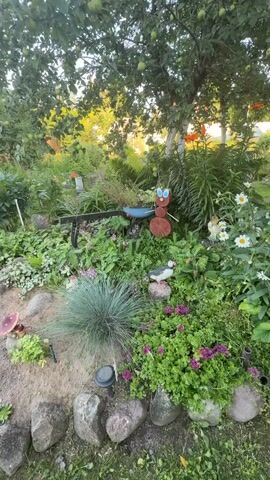 The video pans across a garden decorated with various garden ornaments. In the foreground, there is a small, sandy mound bordered by stones, with a decorative mushroom ornament. To the left, a wooden ant ornament is visible, followed by a blue object that appears to be a miniature wheelbarrow. Further to the right, another wooden ant ornament is placed. The garden is filled with lush greenery, including various flowering plants and trees. The lighting suggests it is daytime, possibly late afternoon. The overall scene depicts a whimsical and colorful garden setting.