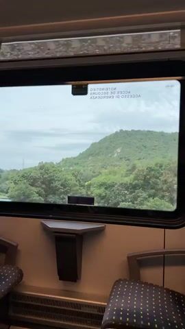 A stationary medium shot from inside a train captures the outside scenery from a window. The train is stopped or moving very slowly as the view outside is relatively still. The window shows a green, tree-covered hillside in the distance under a cloudy sky. In the foreground, through the window, there are trees and some buildings, including what appears to be a swimming pool with blue water. The reflection of the train interior is visible on the window, with inverted text that reads "NOTIENTINETO" and "ACCES DE SECOUR" and "ACCESSO DI EMERGENZA". Below the window, part of the train's beige interior wall and two patterned seats are visible.
