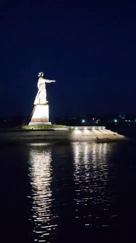 A statue of a woman in a flowing dress stands on a pedestal illuminated by spotlights at night. The statue is situated on a small island or peninsula that extends into a body of dark water. The water reflects the lights from the statue and the surrounding area, creating shimmering patterns. In the background, there is a dark sky with a faint outline of distant lights, possibly from a city or town. The overall scene is serene and atmospheric, emphasizing the grandeur of the illuminated statue against the dark night sky and reflective water.