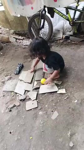 A young child with dark hair, wearing a black shirt and light-colored shorts, is playing on the ground. The child is squatting and interacting with a yellow ball and a toy car, which is black. The child is surrounded by broken pieces of concrete or tile scattered on the dirt ground. In the background, a bicycle with a black frame and yellow accents is leaning against a wall or structure draped with a white fabric that has colorful abstract designs. The environment appears to be outdoors, possibly a yard or alley, with debris and uneven terrain.
