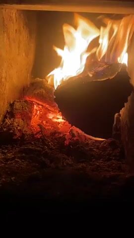 A close-up video shows a roaring fire in a wood-burning stove or furnace. Large logs burn intensely, with bright orange and yellow flames reaching upwards. The interior of the stove is visible, appearing to be made of rough, textured material, possibly brick or stone. Embers glow red at the base of the fire, and a significant amount of ash and burnt material has accumulated on the floor of the stove. The lighting is predominantly from the fire itself, casting a warm, intense glow on the scene. The overall impression is one of intense heat and active combustion.