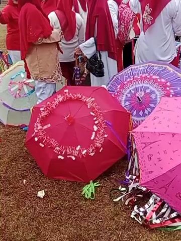 The video shows a ground-level view of several open umbrellas. In the foreground, there is a red umbrella with white and red decorations around its edge. To the right of it is a pink umbrella with a pattern of Eiffel Towers. Behind these two umbrellas, there is a large purple umbrella with a mandala-like pattern. In the background, several people are visible, mostly wearing red and white clothing, some with headscarves. They appear to be standing or walking behind the umbrellas. The ground is covered with what looks like dry leaves or grass. The lighting suggests it is daytime, possibly outdoors.