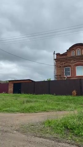 A video captured from a moving vehicle shows a rural scene under a cloudy sky. The camera pans across a dirt road with patchy grass and wildflowers lining the side. On the right side of the road, a series of houses and fences are visible. The houses appear to be made of wood with red roofs and some feature decorative window frames. A dark, possibly black, garage door is set within a brick structure. Power lines stretch across the frame, supported by utility poles. In the distance, a dark-colored car is parked. The overall impression is one of a quiet, possibly somewhat isolated, rural settlement.