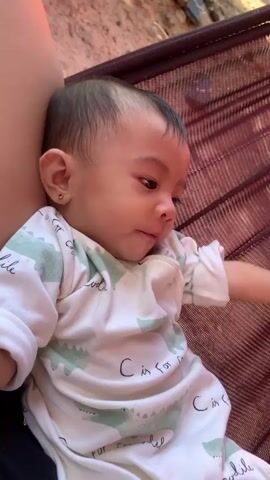 A close-up shot shows a baby lying in a hammock. The baby is wearing a white onesie with a pattern of green crocodiles and the words "C is for crocodile". The baby's head is resting against the side of the hammock, and their right hand is near their face. The background is blurred but appears to be outdoors, with hints of dirt and possibly foliage. The lighting suggests it is daytime. The baby's facial expression is difficult to determine due to the angle and focus.