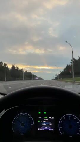 The video shows a first-person perspective of driving on a road. The sky is cloudy with rays of sunlight breaking through. The dashboard of the car is visible, showing speed, battery level, and distance traveled. The road ahead is lined with trees, and there are streetlights on the side of the road. The video appears to have been shot during the daytime, possibly at sunrise or sunset.