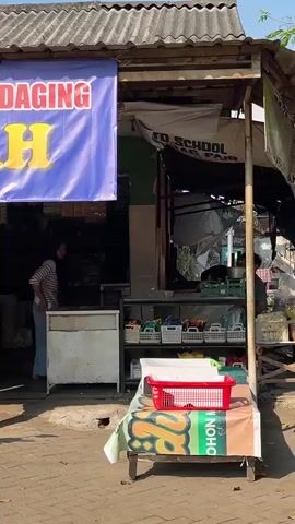 A banner with Indonesian text, partially obscured, hangs over the storefront of a food stall. The visible text includes "NG BAKSO / DAGING" and "DI DAYAH" in red and yellow letters. Underneath the banner, the interior of the stall is visible, showing shelves stocked with items. In front of the stall, several people are present. A young boy in a green shirt stands on the left, next to a motorbike. A young man wearing a blue t-shirt and shorts, carrying a child, stands in the center. A woman in a striped shirt and blue skirt stands further back in the stall's entrance. To the right, a woman in a yellow hijab and blue jeans is interacting with a motorbike, and another person is visible in the background. The ground is paved with bricks, and sunlight suggests it is daytime.
