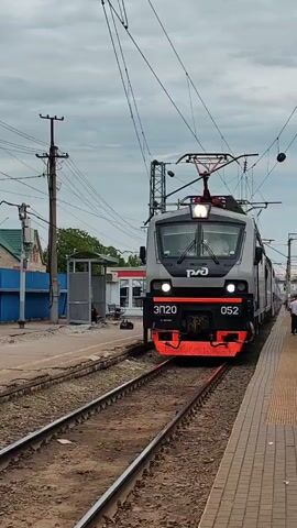 A gray and orange train with the Russian Railways logo is pulling into a station on a cloudy day. The train is marked "EP20 052". It is traveling on a single track, and the platform is on the right-hand side.  A person is standing on the platform near the train. There are power lines overhead. A few buildings are visible in the background.