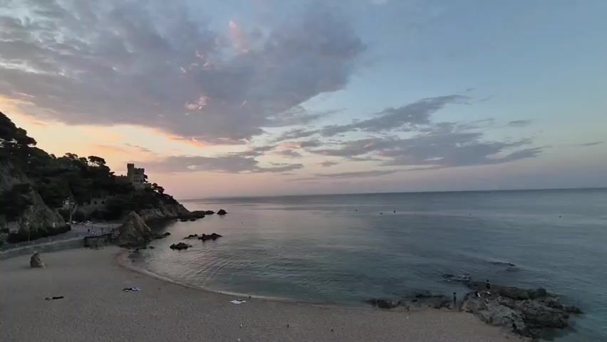 The video captures a wide shot of a serene beach at sunset. The sky is painted with soft hues of pink and orange from the setting sun, with scattered clouds drifting across a pale blue expanse. The calm sea gently laps against a sandy beach, dotted with a few dark rocks in the foreground and along the coastline. On the left side of the frame, a lush green hill rises, crowned by a stone structure that resembles a castle or an old tower. A road runs along the base of the hill, with a few indistinct figures visible walking. On the beach, a few people are scattered, some lying down on towels, and a small group is gathered on the rocks near the water's edge. The overall atmosphere is peaceful and picturesque, showcasing a tranquil coastal landscape as day transitions into night.