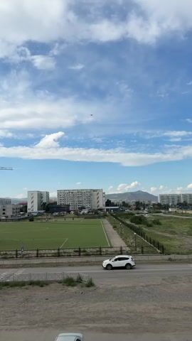 This is a vertical, medium-shot video taken on a cloudy day. The scene is a city with a soccer field in the foreground. In the distance, there are apartment buildings, a construction crane, and hills. A white SUV is driving on the road in front of the soccer field. The sky is blue with white clouds, and a jet is flying high in the sky.