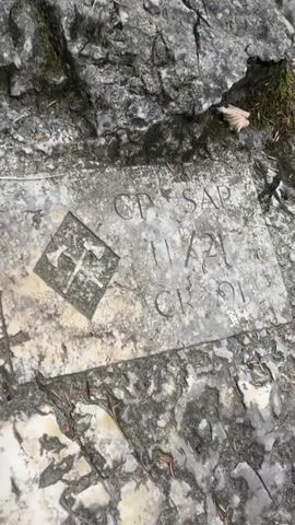 The video begins with a close-up shot of a carved inscription on a rock. The inscription appears to be a date, "11/21", and some letters or symbols. To the left of the inscription is a diamond-shaped emblem with what looks like two crossed axes within it. The rock is set within a larger rocky landscape, with moss and some dry leaves visible on the surface. The camera then pans up and to the right, revealing a scenic view of a waterfall cascading into a pool of water. The surrounding environment consists of large grey rocks, lush green moss, and some foliage. The water is clear and appears to be flowing over a rocky bed. The overall scene suggests a natural, outdoor setting, possibly a hiking trail or a park.