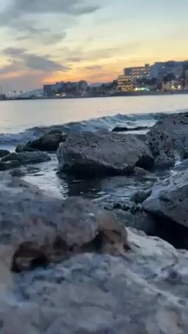 The video shows a coastal scene with rocks in the foreground and a town or city visible in the distance under a twilight sky. The sky has hues of orange and grey. Gentle waves lap against the rocks. The camera appears to be positioned low to the ground. Buildings, trees, and possibly boats are visible in the background.