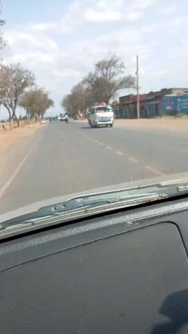 The video, shot from inside a car, shows a view of a straight road. The sky is partly cloudy. Trees line both sides of the road. A few buildings are visible to the right.  There are several motorcycles and vehicles on the road. A person walks across the road from left to right.