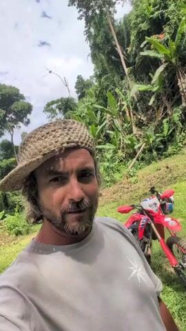 A man wearing a straw hat and a gray t-shirt with a starburst logo is standing in a jungle environment. A red dirt bike is parked behind him. The man is looking directly at the camera with his mouth open. Lush green foliage and trees are visible in the background, with a cloudy sky overhead.
