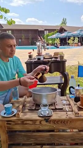 A man is preparing coffee outdoors at a wooden table. He is wearing a teal t-shirt and light shorts. He is holding a wooden coffee grinder in his left hand and is using a metal scoop to put ground coffee into a copper cezve (a Turkish coffee pot). In front of him on the table is a large metal pot, a small glass with a silver base, a spoon, a bottle of water, a moka pot, and a small cup with a saucer. Coffee beans are scattered on the table. In the background, there is a swimming pool, a building with multiple floors, trees, and umbrellas. There are also flags of Romania and another country visible. The sky is clear and blue.