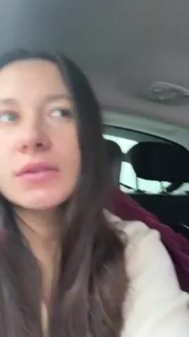A woman with long brown hair and green eyes is seated in a car. She wears a light-colored sweater. The camera is positioned inside the car, focusing on the woman's face. As the video progresses, the camera angle shifts to the right, showing the outside environment through the car window. The landscape is covered in snow, with a line of bare trees bordering the road. The sky appears overcast and white. It is daytime.