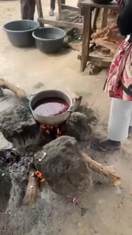 A video captures an outdoor cooking scene. A woman wearing a floral skirt and white pants retrieves a bright orange bucket. The camera then pans to a large silver pot containing a dark liquid and several small pieces of what appear to be cooked meat. This pot sits next to a small green container and a white container, all on the ground. In the background, a pot of vibrant red liquid simmers over an open fire supported by large rocks. Another person stands nearby. The scene suggests communal cooking is taking place.