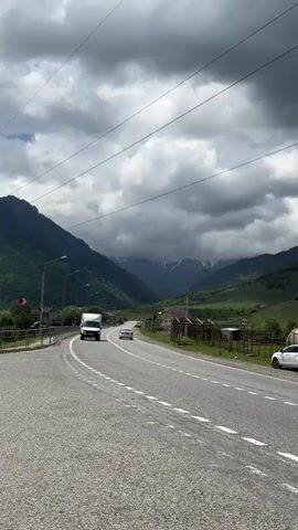 The video shows a view of a road in a mountainous area. A white truck is driving on the road, and several other cars are parked along the side. The mountains are covered in green vegetation, and there are some clouds in the sky. The sky is cloudy, with patches of blue sky visible. There are also some power lines stretching across the frame. A fence runs along the side of the road, and there are some buildings visible in the distance. The road appears to be paved and has lane markings. The overall impression is one of a rural landscape.