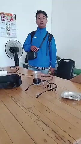 A video recording captures an indoor scene, likely an office or waiting area. The room has white walls, a ceiling fan, and a prominent wooden table in the foreground. The table is cluttered with various items, including a black bag, a cup of dark liquid, a pair of eyeglasses, and some papers.

There are at least three individuals visible. In the first frame, a man wearing a blue jacket and a black crossbody bag stands in the background, looking towards the camera with a neutral expression. He has short dark hair. In subsequent frames, two other men are seen seated at the table. One man, wearing a dark blue shirt and a black baseball cap with glasses, is looking down at his phone. He appears to be touching his chin. The other man, wearing a white t-shirt, is also seated and looking down.

A calendar with visible dates, including "26" and "June", is mounted on the wall. There are also some papers and what appears to be a stack of magazines on the table. The lighting suggests an indoor environment, possibly during the daytime. The video appears to be taken from a handheld device, with some camera movement and slight blurriness at times.