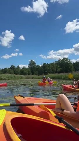 This is a first-person perspective video of a group kayaking on a lake or slow-moving river. The video begins with a close-up of the front of an orange and red kayak, with a yellow paddle resting inside. In the background, several other kayaks are visible, each with one or two people wearing orange life vests. The kayaks are moving through water covered with lily pads and other aquatic vegetation. The shore in the distance is lined with trees and tall grass. The sky is bright blue with fluffy white clouds. The camera pans slightly to show more of the group and the surrounding environment. People in the kayaks are paddling and appear to be interacting with each other. The overall scene depicts a recreational kayaking outing on a sunny day.