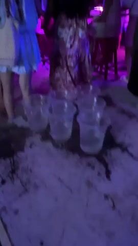 The video shows a lively outdoor party scene at night, likely on a beach or sandy area. Several people are visible, dancing and mingling. The environment is illuminated by various colorful lights, including hanging lanterns in shades of blue, purple, and red, as well as string lights and possibly lights from a nearby establishment. The ground is covered in sand, and there are tables and chairs set up, some with what appear to be drinks or cups on them. The overall atmosphere suggests a festive and energetic gathering.