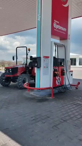 A red tractor is parked next to a fuel pump at a gas station. A man in a red shirt exits the tractor and walks towards the pump. Shortly after, another man joins him near the pump. The gas station has a white canopy and a white building in the background, with other vehicles visible in the distance. The sky is overcast. The fuel pump and overhead canopy prominently display "Aytemiz" and "Optimum" branding, with "Performance ve Tasarruf Maksimum" (Performance and Maximum Savings) partially visible below "Optimum." The setting is a gas station in Acıgöl, Turkey.