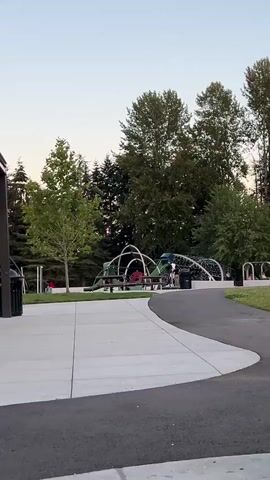A park scene is captured from a low angle. In the foreground, a paved pathway curves from right to left. Beyond the pathway, a concrete plaza features a picnic table and a black trash can. In the middle ground, a vibrant playground structure with green and white arches and netting stands. Several people are visible near the playground, some appearing to be children. To the left of the playground, a lone tree with green foliage is prominent, and behind it, a line of dark green trees forms a backdrop. Further to the right, a row of white swings and more trees are visible. The sky above is a pale, hazy blue. The overall impression is of a peaceful outdoor recreational space during daylight hours, possibly late afternoon given the soft lighting.