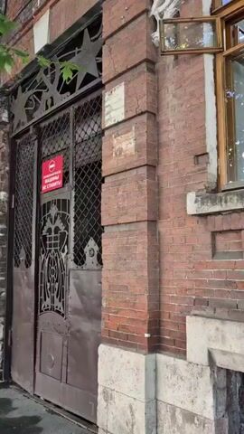 The video begins showing a close-up of a red brick building's facade, featuring an ornate metal gate on the left side, a partially opened wooden window above it with a white decorative element next to it, and a darker brick wall with a white stone foundation below. As the camera pans right, the building continues, revealing more windows with wooden frames and decorative white elements above them. A drainpipe runs vertically down the brick wall, and leafy green branches from a tree overhang the top of the building. The camera then pans further right to show a sidewalk made of gray interlocking pavers and a street visible in the background with a pedestrian crossing sign and a few people walking. The video appears to be filmed outdoors during the daytime, likely in a city or town environment.
