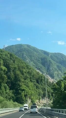 The video shows a view from inside a vehicle moving along a highway. The highway is surrounded by lush green mountains under a clear blue sky with a few white clouds. Several cars are visible on the highway, including a white van, a white SUV, and a dark-colored SUV in the foreground. A tall street lamp is positioned on the right side of the road. The camera angle suggests a moving perspective, likely from the passenger seat or dashboard. The overall scene is one of travel through a scenic, mountainous landscape.