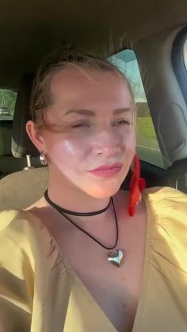 A woman with blonde hair pulled back, wearing a yellow top, a red scarf tied around her head, a black choker necklace with a heart pendant, and large hoop earrings, is filmed from the chest up inside a car. She is looking to her right, with sunlight reflecting off her face. The background shows a road and trees through the car window. The video consists of a single shot.