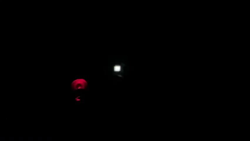 The video is a dark, low-light shot of what appears to be a computer setup in a room. The primary subject is a computer tower with several illuminated fans, displaying a vibrant, multicolored RGB lighting effect that cycles through various colors like red, green, blue, and purple. To the left of the computer, there is a smaller, red illuminated object, possibly another fan or a decorative light. A small, rectangular screen or indicator light is also visible on the computer tower. The overall setting is dark, suggesting it is nighttime or the room is dimly lit. The focus is on the glowing components of the computer, creating a visually striking effect against the darkness. There are no people visible in the video, and no discernible text. The location context provided is Ардонь, Russia, but there are no visual cues within the video to confirm this specific location. The atmosphere conveyed is one of quiet intensity, with the pulsating lights being the main source of visual interest.