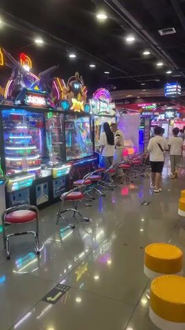 The video shows an indoor arcade with multiple rows of brightly lit game machines. People are scattered throughout the arcade, playing games, watching others play, or walking around. In the foreground, there are several rows of red and chrome stools in front of some of the game machines. The floor is shiny and reflects the lights from the machines. In the background, more arcade games are visible, along with a variety of people of different ages. Some people are standing or sitting near the machines, while others are walking through the aisles. The overall atmosphere appears to be lively and engaging.
