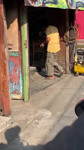 A person wearing a yellow shirt and cargo pants bends over in a workshop. Stacks of tires are visible in the background, along with tools and equipment. The person appears to be working on something on the floor. Sunlight streams into the workshop, casting shadows.