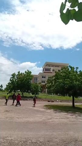 The video begins with a view of a paved area with some greenery and buildings in the background. Several people are visible walking in different directions. As the video progresses, the camera pans and shows more of the surroundings, including trees, more buildings, and a cloudy sky. Some individuals are seen walking together, while others are walking alone. The scene appears to be an outdoor public space, possibly a campus or park, with people engaged in everyday activities like walking. The camera then shifts focus to show a group of people walking away from the camera down a paved path lined with trees. The video concludes with another wide shot of the environment, showing people walking in the distance and the overall setting.