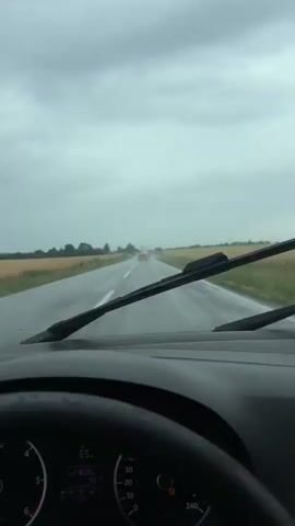 The video shows a first-person view from inside a car driving on a wet road under a cloudy sky. The windshield wipers are active, clearing rain from the glass. The car is moving along a rural road flanked by fields of dry grass or crops. In the distance, other vehicles can be seen on the road. The dashboard and steering wheel are visible in the foreground, indicating the driver's perspective. The weather appears to be overcast and rainy, contributing to the wet road conditions.