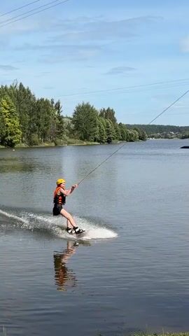 This video shows a person wakeboarding on a lake. They are wearing a helmet and a life vest. The wakeboarder is being pulled by a rope attached to a boat or cable system. There are trees and a shoreline in the background. The video also shows a dock with a person standing on it and some wakeboards. There are also some people in boats on the lake. The sky is blue with some clouds.