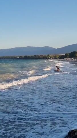 The video shows a scenic beach with calm waters and distant mountains. Two people are seen playing in the shallow water near the shore, with gentle waves lapping around them. The sun is shining, casting a warm glow on the scene, and the sky is a clear blue. The beach itself appears to be a pebble beach, with a few scattered trees and buildings visible in the background. The overall atmosphere is peaceful and serene.