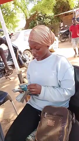 A young woman with a brown headscarf is sitting on a chair in an outdoor setting. She is wearing a light blue long-sleeved shirt and black pants.  In her hands, she holds a stack of banknotes, which she appears to be counting. A brown satchel bag sits beside her on the chair. A table with a calculator on it is visible to her left. In the background, there are people walking around, parked motorcycles, and a white car. There are also trees, suggesting a public area like a market or a street. The camera is stable, focused on the woman counting money.