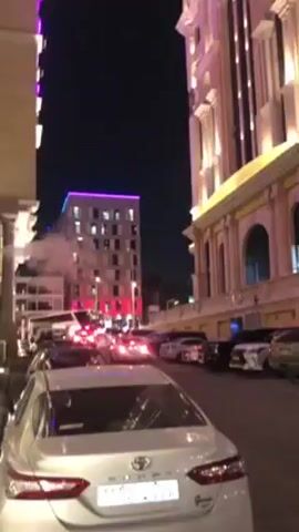 The video shows a street scene at night. The camera pans upwards, revealing several buildings illuminated with lights. Cars are parked along the street, and there is some traffic. In the distance, a tall skyscraper is visible, highlighted by blue laser beams. The overall setting appears to be an urban area with a combination of modern and traditional architecture.