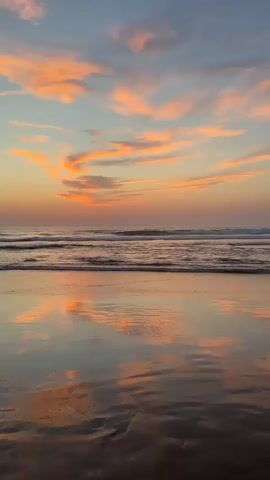 The video shows a scenic view of the ocean at sunset. The sky is filled with vibrant orange and pink clouds, reflected in the calm water and wet sand of the beach. Gentle waves roll towards the shore, creating a serene and peaceful atmosphere. No people or specific objects are visible, emphasizing the natural beauty of the coastal landscape.