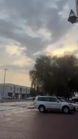 The video shows a residential area after rainfall, with wet roads and puddles. The sky is overcast with clouds, but there's a glow of sunlight filtering through them. Various cars are parked on the street and in front of the houses. The houses are mostly two-story buildings with similar architectural styles. A few trees are visible.
