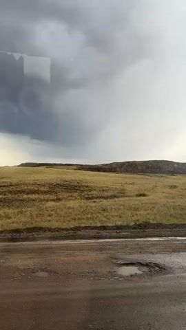This media file captures a view of a rural landscape, likely taken from a vehicle. The primary subject is a vast, open field covered in dry, yellowish-brown grass, with patches of green visible closer to the foreground. The field is sloped, leading up to a distant, gently rolling hillside or mesa that forms the horizon. The sky above is dramatic, with heavy, dark grey storm clouds dominating the left side and a brighter, lighter grey or overcast sky to the right. There are visible streaks or shafts of light breaking through the clouds in the distance, suggesting the storm may be moving or breaking.

In the immediate foreground, there is a dirt road that appears wet and muddy, with several puddles reflecting the overcast sky. The road is textured with gravel and dirt, showing signs of wear and tear. The overall scene evokes a sense of rugged, natural beauty, with the dramatic weather contributing to a somewhat somber or atmospheric mood.

The location cues provided place this scene in Karagayl, Kazakhstan. The time of day appears to be late afternoon or early evening, given the lighting conditions and the overcast sky. The weather is clearly inclement, with signs of recent or ongoing rain. There are no people or specific objects in the scene, other than the natural landscape and the road. No visible text is present within the media file. The overall impression is of a remote, open terrain experiencing unsettled weather.