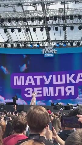 A woman in a light pink dress stands on a stage performing for a large crowd. She is holding a microphone and appears to be singing. Behind her, a large screen displays the text "МАТУШКА ЗЕМЛЯ" in large purple letters. Above the screen, a banner reads "VK Fest 2026". To the right of the singer, a person in a black outfit is filming her with a video camera. The crowd in front of the stage is holding up phones to record the performance. The environment appears to be an outdoor festival, with stage lighting rigs visible overhead.