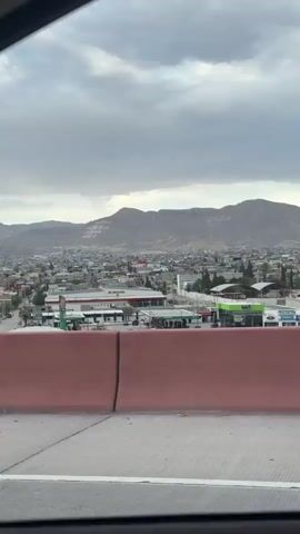 The video begins by showing a panoramic view of a city with mountains in the background. The sky is cloudy. As the video progresses, the camera pans to the right, revealing more of the city, including buildings, roads, and vehicles. A black SUV is seen driving on the road, followed by other vehicles. The video then shows a fence and some buildings in the distance. The sky is still cloudy, but there are also patches of blue sky visible. The video ends with a view of the city from a bridge.