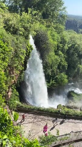 A video shows a wide waterfall from an elevated viewpoint with people visible below. The waterfall itself is large and powerful, surrounded by lush green trees and vegetation. A paved walkway runs along the edge of the viewpoint, separated from the drop-off by a low metal railing. Several people are walking and standing on the walkway, some appearing to observe the waterfall. Beyond the waterfall, the landscape stretches out into the distance, featuring more trees, rolling hills, and a clear blue sky. The overall scene is bright and sunny.