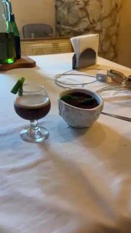 The scene is a close-up shot of a table setting, probably in a restaurant, with a white tablecloth. On the table, there's a small glass of dark liquid garnished with a lime wedge, and a white, textured bowl filled with a dark liquid and garnished with green leaves. Behind these items are bottles, a tissue holder, phone chargers, and sunglasses. A hand wearing a beaded bracelet walks its fingers across the table towards the bowl. The background features a stone wall and a partially visible cane-back chair. The lighting is warm.