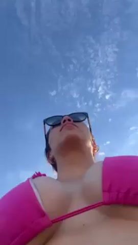 A woman in a pink bikini and sunglasses stands outside on a sunny day, holding the camera and filming herself. She has tattoos on her left arm and is smiling. Several people walk in the background. The sky is blue with scattered clouds.