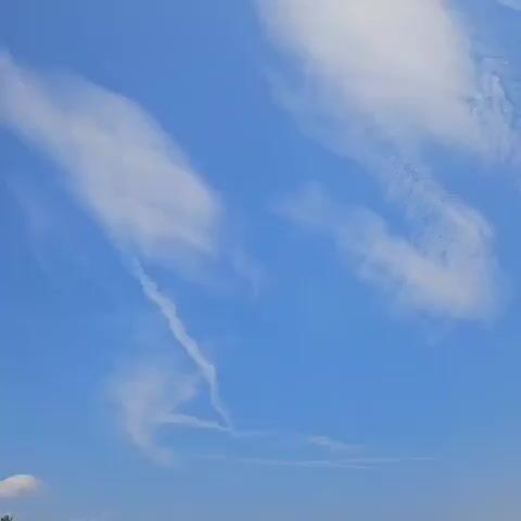 A video shows a blue sky with white clouds. The sun is visible and bright, with rays extending outwards. Several wispy clouds and contrails are also present in the sky.